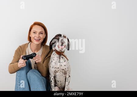 KIEW, UKRAINE - 16. DEZEMBER 2022: Lächelnde rothaarige Frau spielt Videospiel in der Nähe des dalmatinischen Hundes Stockfoto