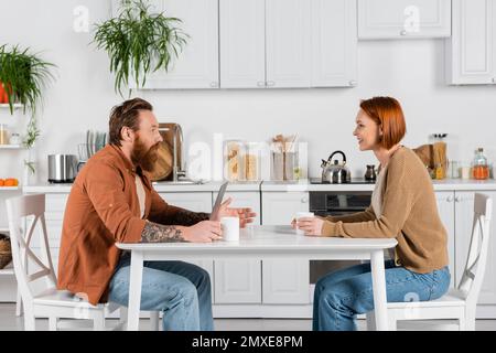 Seitenansicht eines bärtigen Mannes, der mit einer rothaarigen Frau in der Nähe des Laptops und der Tassen in der Küche spricht, Stockbild Stockfoto