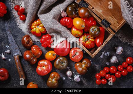 Viele verschiedene Arten, Formen und Größen von Tomaten in einer alten Holzkiste und auf einer dunklen Oberfläche, flach liegend, Draufsicht. Erntekonzept Stockfoto