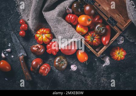 Viele verschiedene Arten, Formen und Größen von Tomaten in einer alten Holzkiste und auf einer dunklen Oberfläche, flach liegend, Draufsicht. Erntekonzept Stockfoto