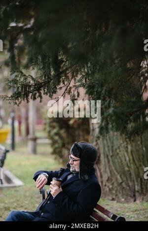 Ein älterer Mann genießt einen kalten Wintermorgen in der Stadt. Ein Mann sitzt auf einer Bank auf dem Platz, das Konzept von Frieden und Vitalität. Stockfoto