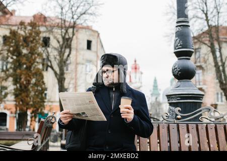 Ein älterer Mann genießt einen kalten Wintermorgen in der Stadt. Ein Mann sitzt auf einer Bank auf dem Platz, liest Zeitung und trinkt Kaffee. Stockfoto