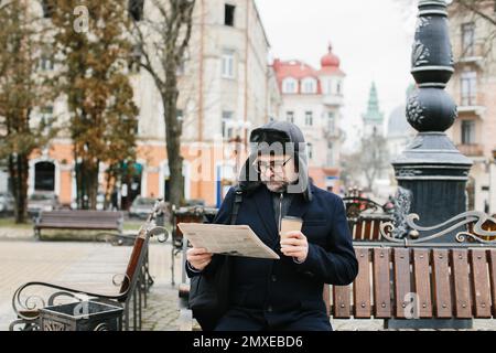 Ein älterer Mann genießt einen kalten Wintermorgen in der Stadt. Ein Mann sitzt auf einer Bank auf dem Platz, liest Zeitung und trinkt Kaffee. Stockfoto