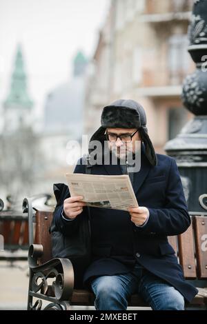 Ein älterer Mann genießt einen kalten Wintermorgen in der Stadt. Ein Mann sitzt auf einer Bank auf dem Platz, liest Zeitung und trinkt Kaffee. Stockfoto