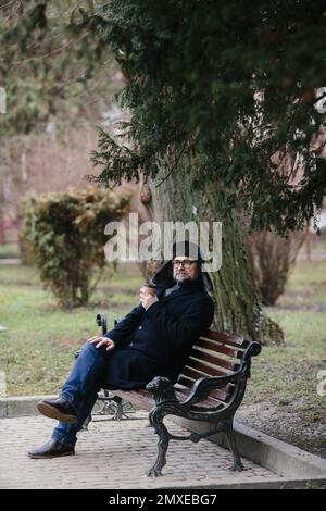 Ein älterer Mann genießt einen kalten Wintermorgen in der Stadt. Ein Mann sitzt auf einer Bank auf dem Platz, das Konzept von Frieden und Vitalität. Stockfoto