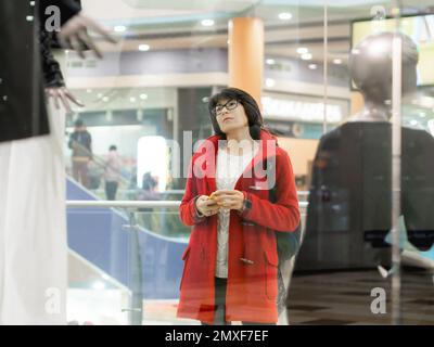 Die Frau schaut auf Dummys im Bekleidungsgeschäft. Lässiges Outfit, das an Schaufensterpuppen hängt. Blick durch transparentes Schaufenster. Einkaufen im Einkaufszentrum. Stockfoto