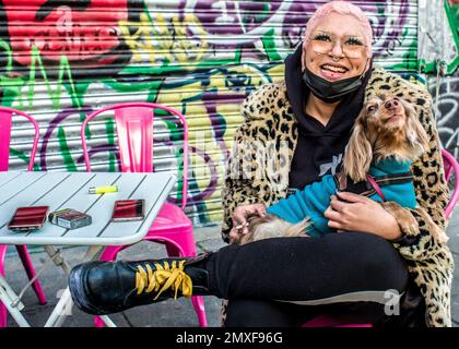 Weibliche Straßenhändlerin mit rosafarbenen Haaren trägt einen Mantel mit Leopardenmuster, Doc Marten Stiefel hält einen Hund, lächelt in die Kamera. Camden Markt Stockfoto