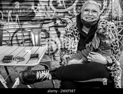 Weibliche Straßenhändlerin mit rosafarbenen Haaren trägt einen Mantel mit Leopardenmuster, Doc Marten Stiefel hält einen Hund, lächelt in die Kamera. Camden Markt Stockfoto