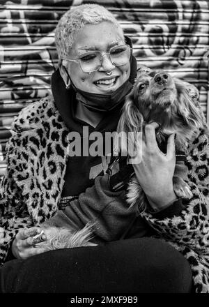 Weibliche Straßenhändlerin mit pinkfarbenen Haaren trägt einen Mantel mit Leopardenmuster, Doc Marten Stiefel hält einen Hund und lächelt in die Kamera. Camden Markt Stockfoto