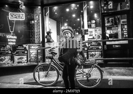 Eine Frau mit Wollmütze und Radhelm starrt direkt in die Kameralinse und wartet neben dem Fahrrad Stockfoto