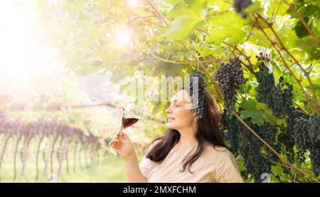 Wunderschöne Frau träumt von Rotwein und genießt den Sommeraufenthalt in Weinbergen an einem schönen sonnigen Tag. Eine Frau, die Rotwein trinkt. Erntesaison. Hochwertiges Foto Stockfoto