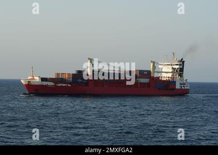 Beladenes Containerschiff mit rotem Rumpf und zwei Ladekränen nähert sich der Ankerposition. Stockfoto