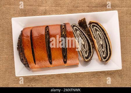 Eine appetitliche Mohnrolle auf weißer Keramikplatte auf Jutetuch, Makro, Draufsicht. Stockfoto