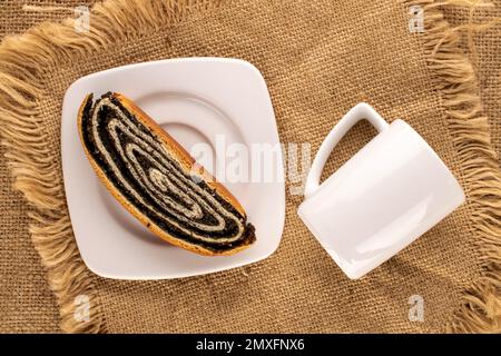 Ein Stück schmackhafte Mohnrolle auf weißer Keramikplatte mit Tasse auf Jutetuch, Makro, Draufsicht. Stockfoto