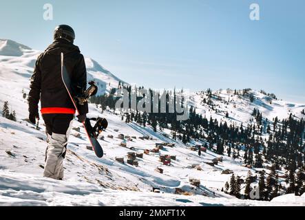 Ein Tourist im Skianzug mit Snowboard in den Händen wandert auf einer schneebedeckten Piste vor dem Hintergrund einer wunderschönen Berglandschaft Stockfoto