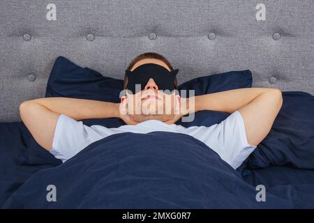 Draufsicht Foto eines jungen Mannes mit Schlafmaske, fröhlichem positivem Lächeln, Schlafdecke, Decke, Kleidung, Schlafanzug Stockfoto