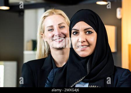Lächelnde Geschäftsfrauen mit unterschiedlichen ethnischen Zugehörigkeiten und Kulturen, die in einem modernen Büro vor die Kamera schauen - zwei elegante Freundinnen, eine davon trägt Ty Stockfoto