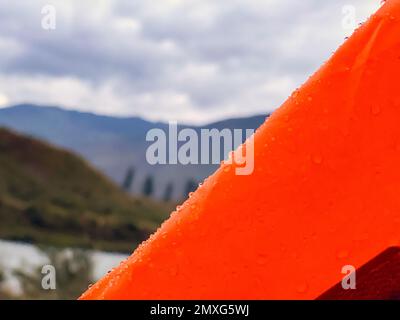 Am Rand der Markise des orangefarbenen Zelts liegen am Abend in Altai Regentropfen vor dem Hintergrund des Flusses und der Berge. Stockfoto