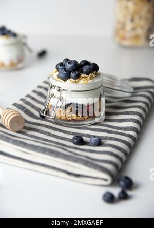 Haferflocken zum Frühstück mit Obst in einem Glasgefäß aus griechischem Joghurt und Müsli garniert mit frischen Blaubeeren auf gestreiftem Tuch mit Honiglöffel, Gesundheit Stockfoto