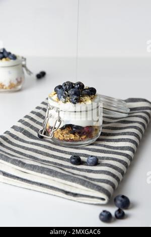 Haferflocken zum Frühstück mit Obst in einem Glasgefäß aus griechischem Joghurt und Müsli garniert mit frischen Blaubeeren auf gestreiftem Tuch mit Honiglöffel, Gesundheit Stockfoto