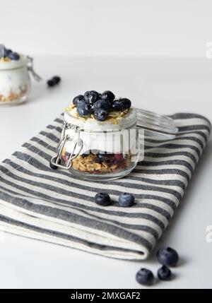 Haferflocken zum Frühstück mit Obst in einem Glasgefäß aus griechischem Joghurt und Müsli garniert mit frischen Blaubeeren auf gestreiftem Tuch mit Honiglöffel, Gesundheit Stockfoto