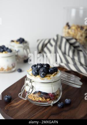 Haferflocken zum Frühstück mit Obst in einem Glasgefäß aus griechischem Joghurt und Müsli garniert mit frischen Blaubeeren auf gestreiftem Tuch mit Honiglöffel, Gesundheit Stockfoto