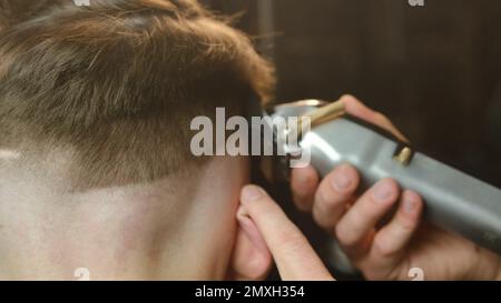 Schließen Sie das Friseur- und Haarschneiden von Männern mit einem Haarschneider in einem Friseursalon oder Friseursalon. Friseurservice in einem modernen Friseursalon mit dunklem Schlüssel Stockfoto