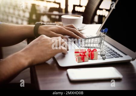 Ein Mann kauft online mit einem Laptop, einem kleinen Wagen und einer Schachtel auf dem Computer ein Stockfoto