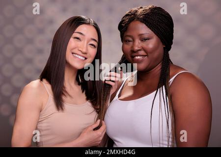 Asiatische Frau berührt afroamerikanische Dame lange Zöpfe Haarschnitt. Zwei hübsche junge, vielfältige Haarpflegemodelle, die zusammen posieren, in die Kamera schauen und lächeln Stockfoto