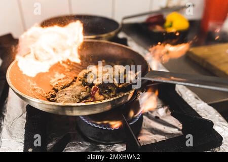 Schweinebraten mit Pilzen in der brennenden Bratpfanne. Speisezubereitung in der Küche des Restaurants. Horizontale Indoor-Aufnahme. Hochwertiges Foto Stockfoto