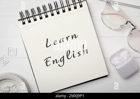 Notizbuch mit Text Lernen Sie Englisch, Brille und Ohrhörer auf einem weißen Holztisch, flach liegend Stockfoto