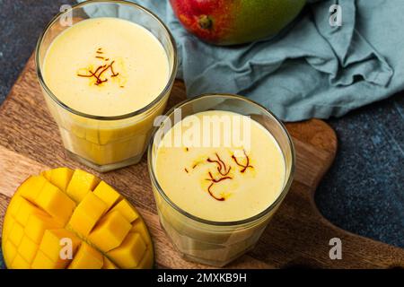 Gesundes indisches Ayurveda trinken Mango in zwei Gläsern auf einem rustikalen Betontisch mit frischer, reifer Mango, gelbes Mischgetränk aus Mango fru Stockfoto