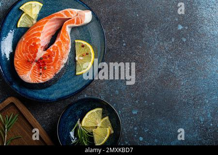 Ungekochter roher Fisch Lachs Steak von oben auf Teller rustikaler dunkler Betonstein Hintergrund mit Rosmarin, Zitronenspalten und Gewürzen, Delikatesse gesund Stockfoto