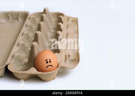 Ein einzelnes Hühnerei mit traurigem Gesicht in einem offenen hellbraunen Karton, Dutzend Eierkarton, links vom Rahmen auf einer sauberen weißen Oberfläche Stockfoto