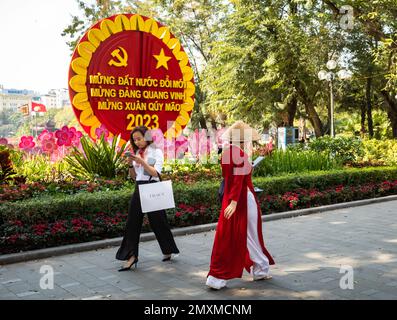 Eine Frau in traditionellem AO dai mit einem konischen Hut sieht ihr Smartphone an, während eine kostspielig gekleidete Frau in die entgegengesetzte Richtung in die Vergangenheit geht Stockfoto