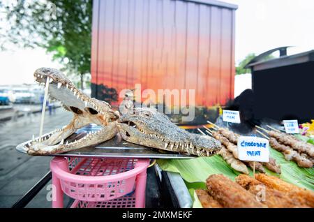 Ein Krokodilfleisch, Straßenverkäufer zeigt zwei getrocknete Krokodilköpfe, Münder auf Affen, mit scharfen Zähnen, neben verschiedenen Kebabs und anderen Fleischzubereitungen, Stockfoto