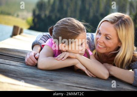 Sie kichert über Frauengespräche. Eine Mutter und eine Tochter, die auf einem Pier an einem See liegen. Stockfoto