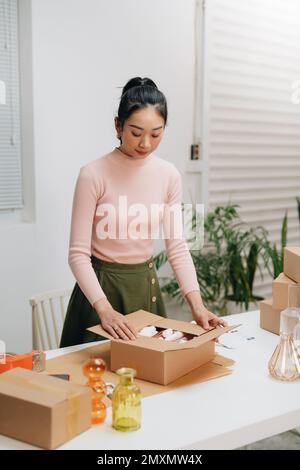 Junge, schöne, glückliche asiatische Geschäftsfrau, die online ein KMU-Notebook benutzt, erhält eine Bestellung von einer Kundin mit Paketverpackung in ihrem Startup-Haus Stockfoto