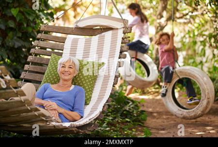 Die Affen beschäftigen. Porträt einer Seniorin, die in einer Hängematte im Garten liegt, während ihre Enkel im Hintergrund spielen. Stockfoto