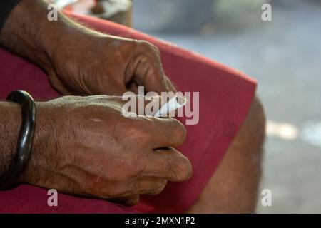 Eine kräftige Hand mit brauner Haut, die ein Armband aus pflanzlichen Wurzeln trägt, rollt Tabak vor einem Hintergrund mit roten Shorts. Stockfoto