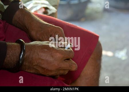 Eine kräftige Hand mit brauner Haut, die ein Armband aus pflanzlichen Wurzeln trägt, rollt Tabak vor einem Hintergrund mit roten Shorts. Stockfoto