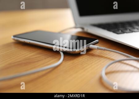 Modernes Smartphone zum Aufladen über Laptop auf Holztisch, Nahaufnahme Stockfoto