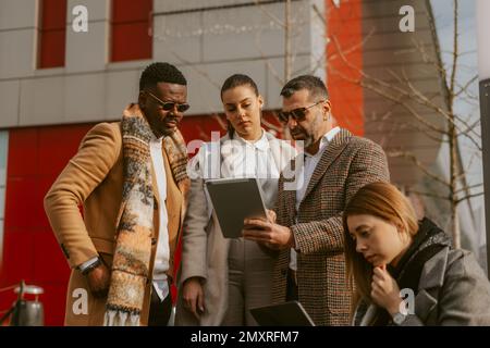 Geschäftsleute denken und schauen sich das Tablet an, während einer von ihnen sitzt, während die anderen drei in der Nähe des Einkaufszentrums stehen Stockfoto