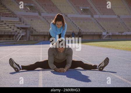 Ein gutaussehender Junge dehnt seinen Körper. Seine Freundin hilft ihm, sie drückt seinen Rücken auf den Boden Stockfoto