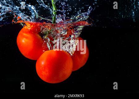 Frische rote Tomate, die ins Wasser fällt, mit Wasserspritzer und Luftblasen auf schwarzem Hintergrund. Stockfoto