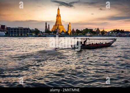 In der Dämmerung gleitet die Silhouette eines thailändischen Langschwanzboots vorbei an den goldenen Pagoden der Tempelanlage, während sie auf den klaren Sonnenuntergang zeigen Stockfoto