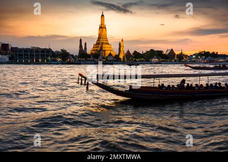 In der Dämmerung gleitet die Silhouette eines thailändischen Langschwanzboots vorbei an den goldenen Pagoden der Tempelanlage, während sie auf den klaren Sonnenuntergang zeigen Stockfoto