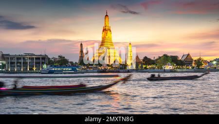 In der Dämmerung gleitet die Silhouette eines thailändischen Langschwanzboots vorbei an den goldenen Pagoden der Tempelanlage, während sie auf den klaren Sonnenuntergang zeigen Stockfoto