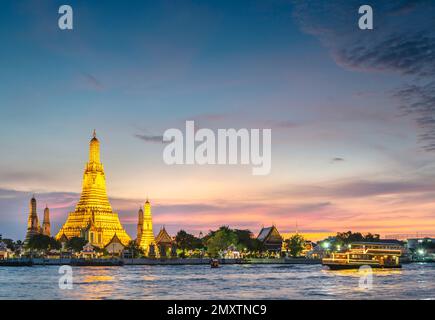 In der Dämmerung gleitet die Silhouette eines thailändischen Langschwanzboots vorbei an den goldenen Pagoden der Tempelanlage, während sie auf den klaren Sonnenuntergang zeigen Stockfoto