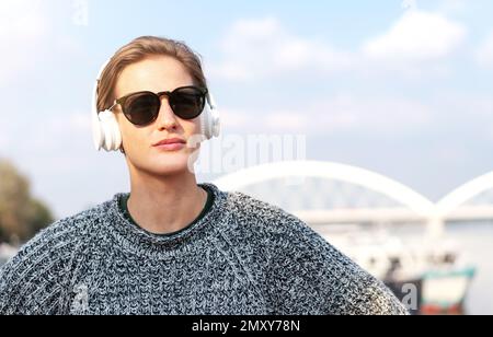 Porträt stilvolle Frau mit kurzen Haaren in Kopfhörern und Sonnenbrillen im Freien gegen Brücke und Himmel. Stockfoto